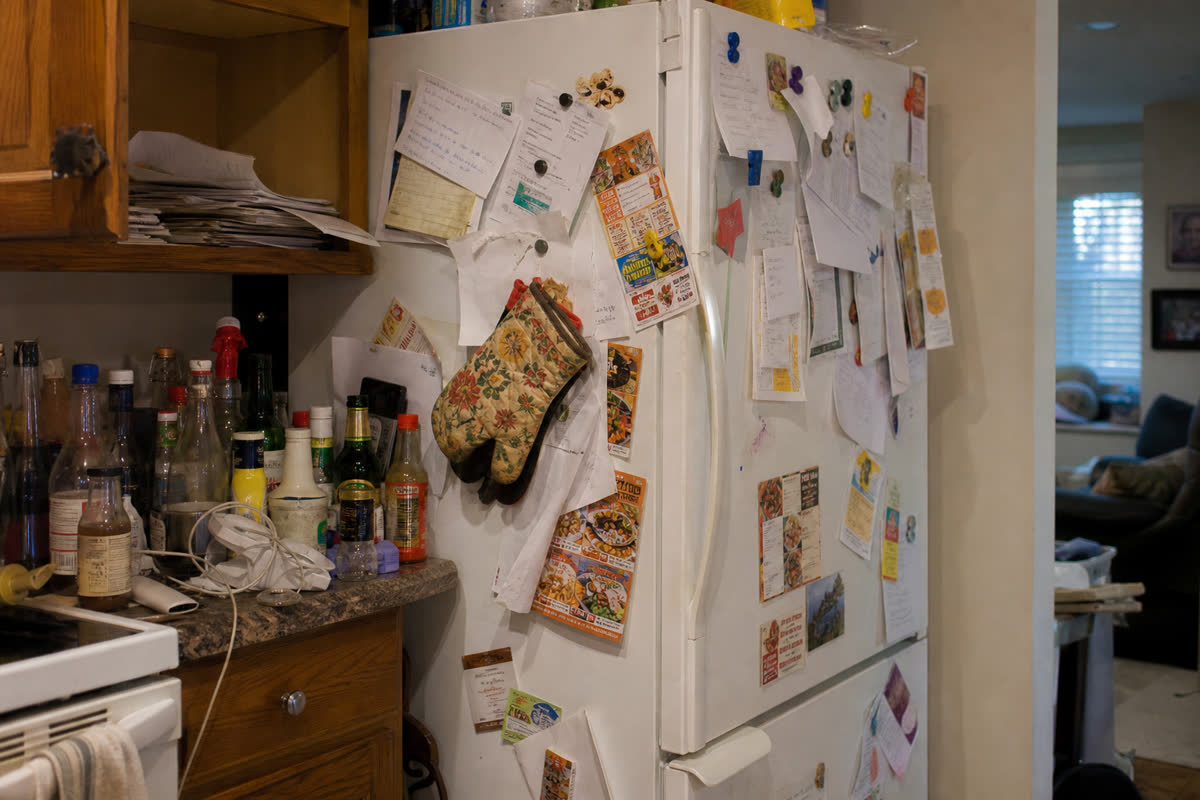 A cluttered kitchen refrigerator covered in papers and bottles on the counter